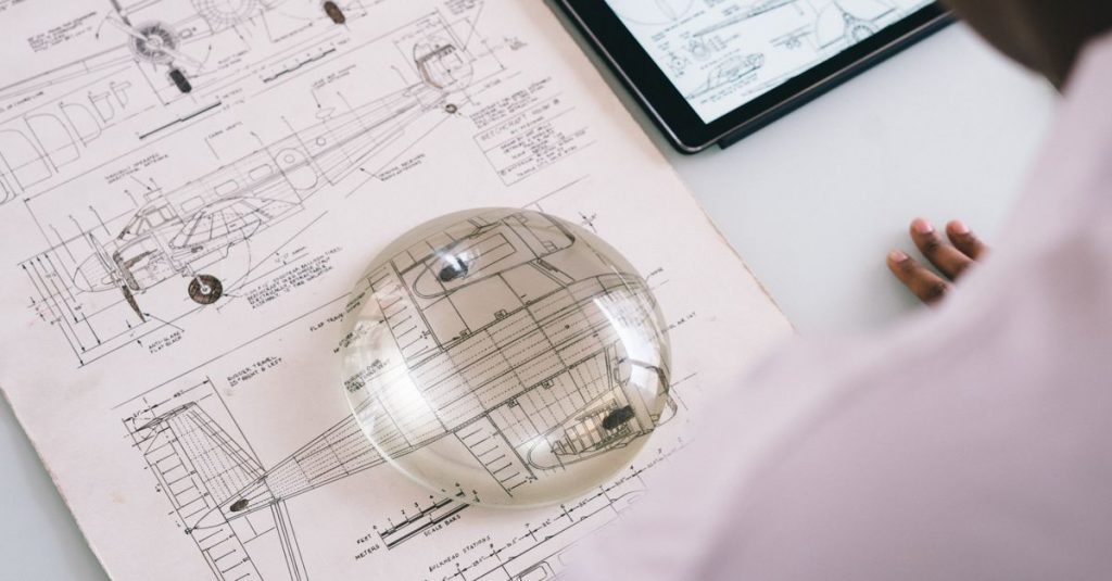Crop unrecognizable mechanic holding magnifying glass on paper with airplane drawing while working on project on tablet in aircraft factory